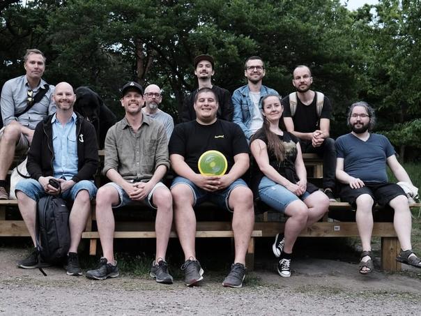 Tretton personer sitter på träbänkar utomhus framför skogen, en av dem håller en frisbee