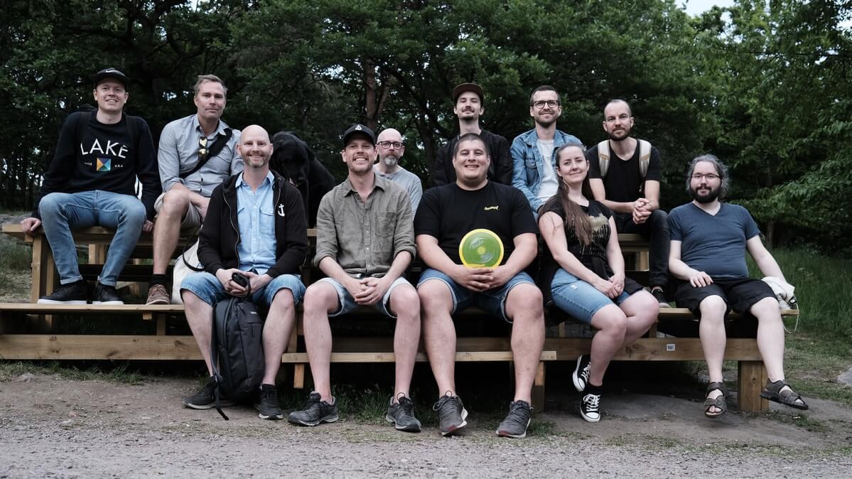 Tretton personer sitter på träbänkar utomhus framför skogen, en av dem håller en frisbee