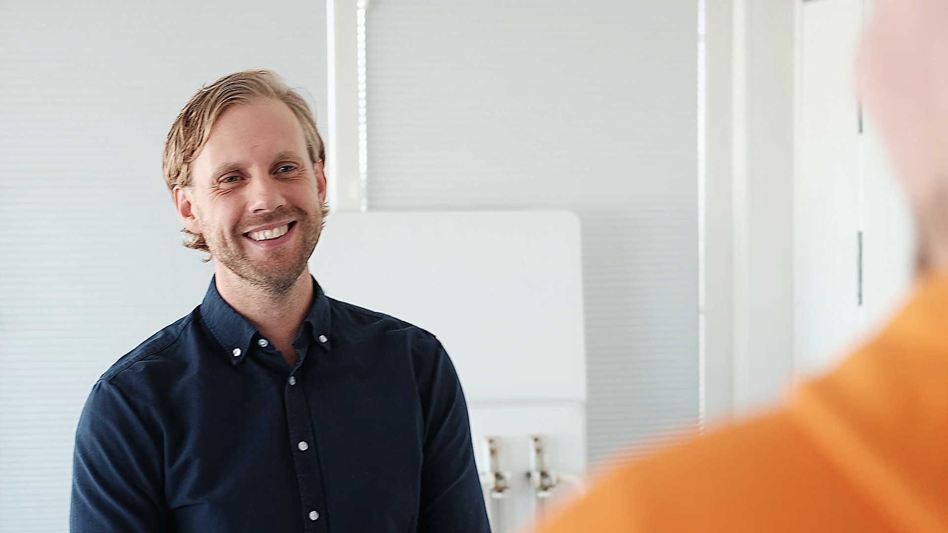 Leende man i mörkblå skjorta som står i ett kontor och pratar med en kollega